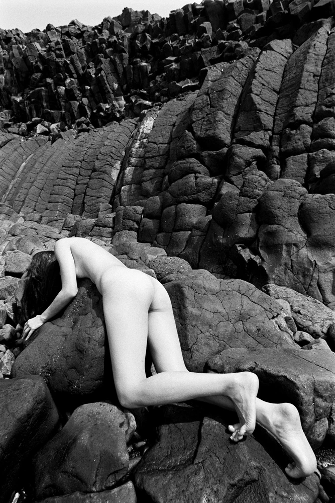 Black and white nude study of a female figure posed among dark volcanic rock, emphasising sculptural form through tonal contrast.