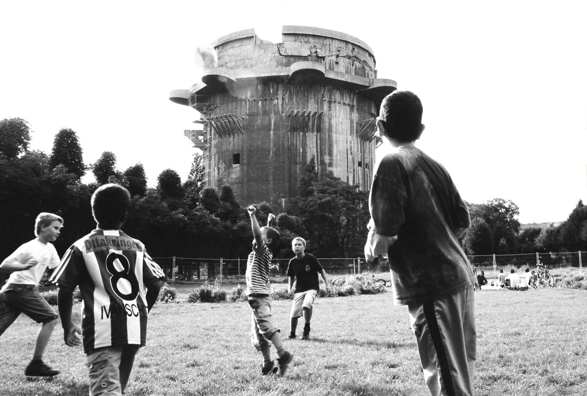 Children playing beneath a towering industrial concrete structure in a black and white urban landscape, contrasting youthful energy with monumental architecture.
