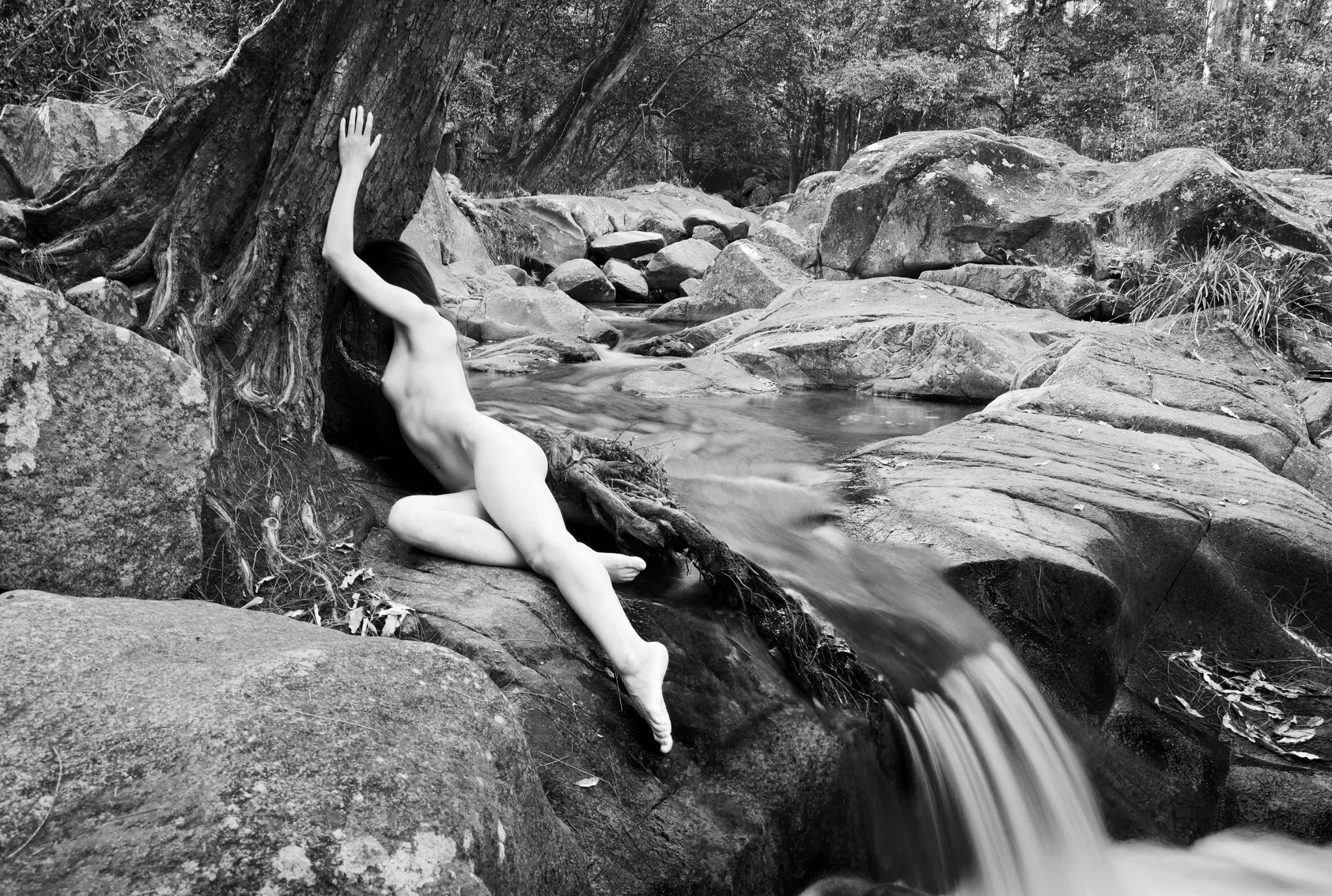 Black and white nude study of a female figure lying on rocks beside a gentle cascade, where the human form blends with the natural landscape.