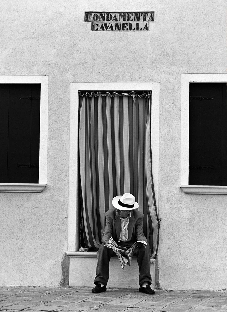 Man in a white hat reading a newspaper beneath the FondamentaCavanella sign in Venice, framed by striped curtains and minimalist architecture in black and white.