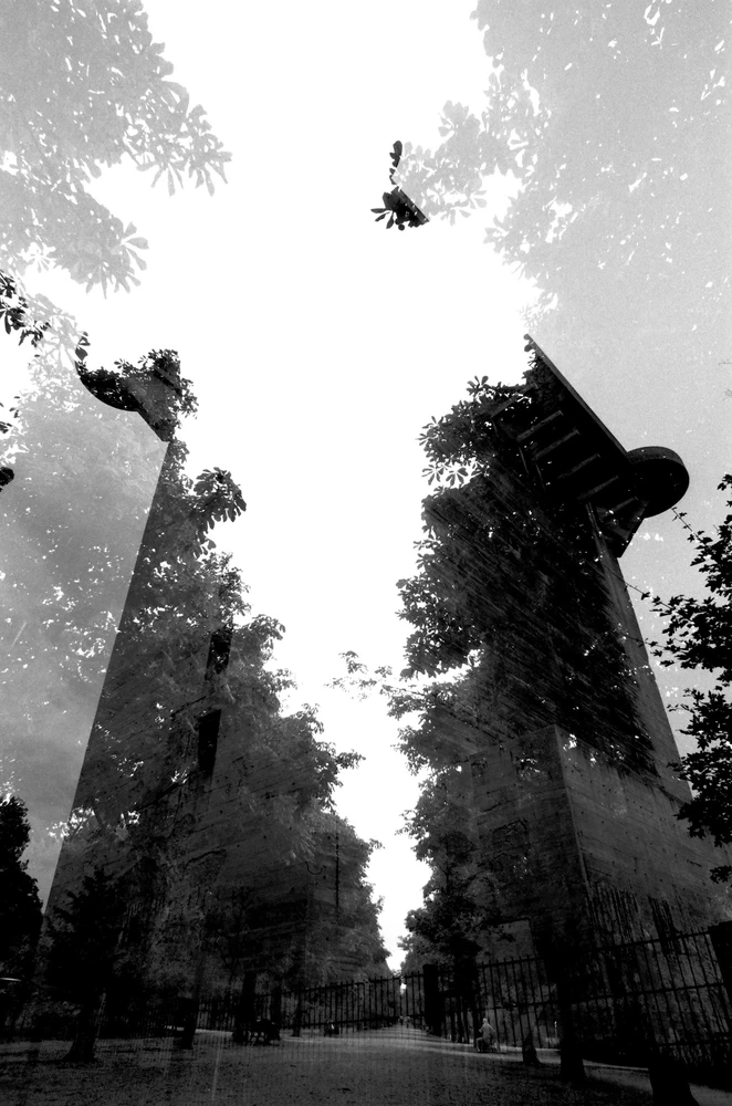 Black and white multiple exposure photograph of a brutalist tower merging with trees and foliage, suggesting architecture being reclaimed by nature.