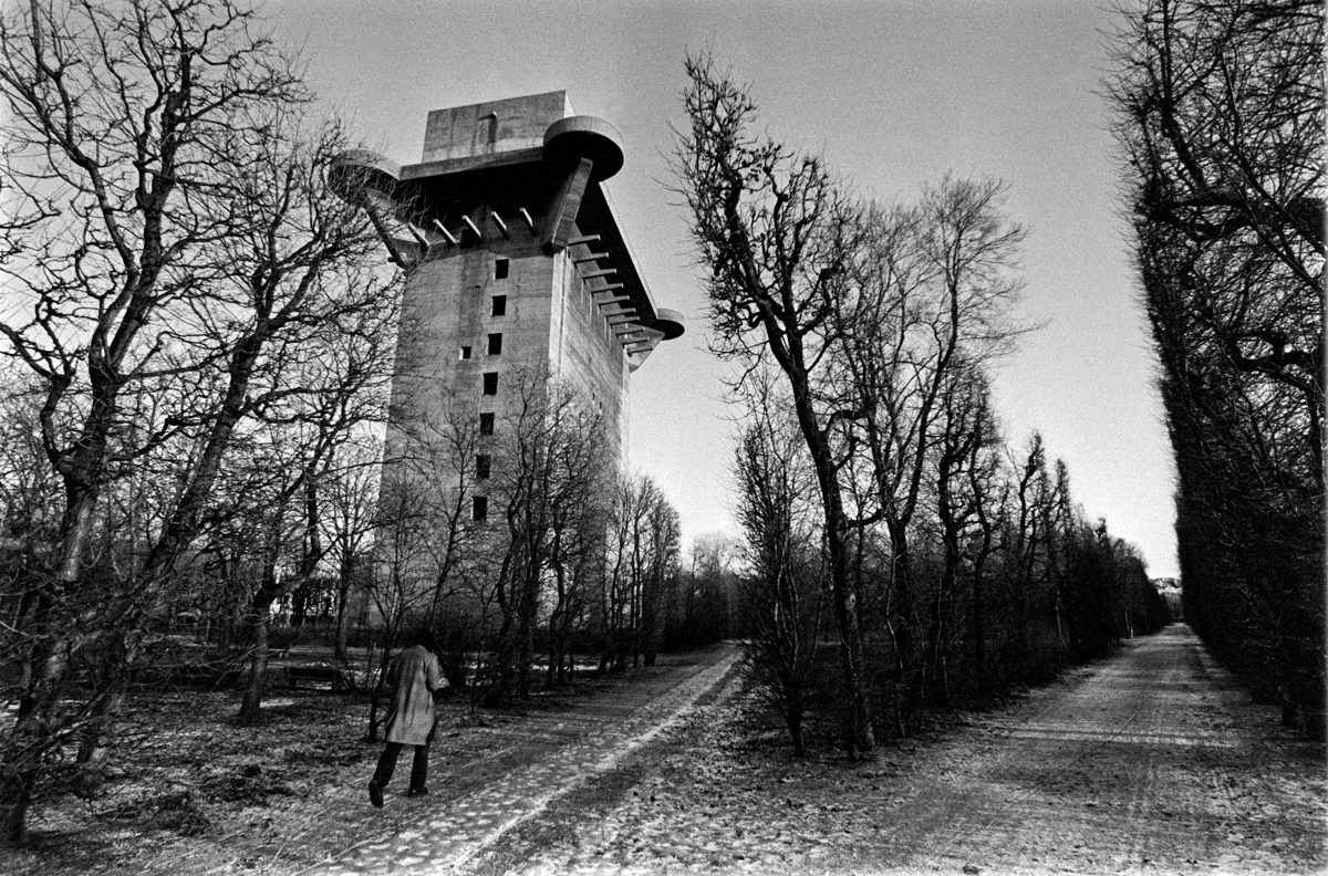 Black and white brutalist architecture photograph of a towering concrete structure framed by bare winter trees and a solitary figure