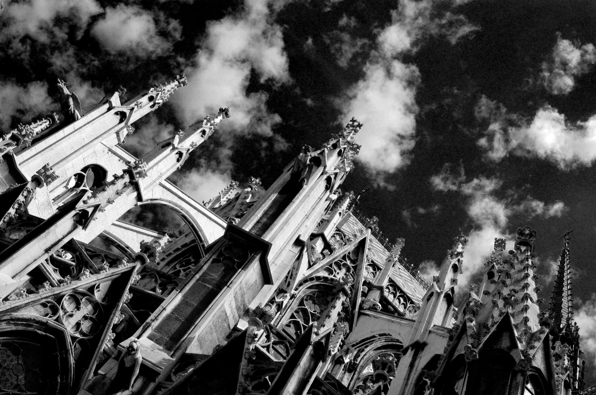 Black and white photograph of Gothic cathedral spires rising into dramatic clouds, emphasising ornate architectural detail and soaring perspective.
