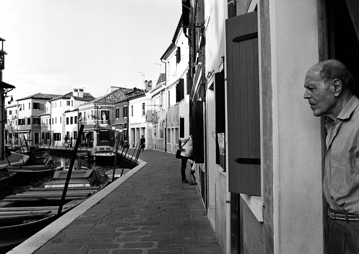 Elderly man standing in a doorway overlooking a quiet Venetian canal in black and white.