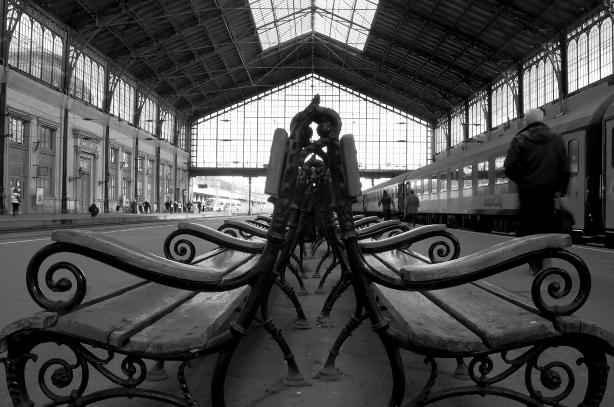 Black and white fine art photograph of ornate benches inside Budapest Nyugati Train Station, featuring symmetrical ironwork, striking station architecture, and blurred travellers in a quiet geometric composition.