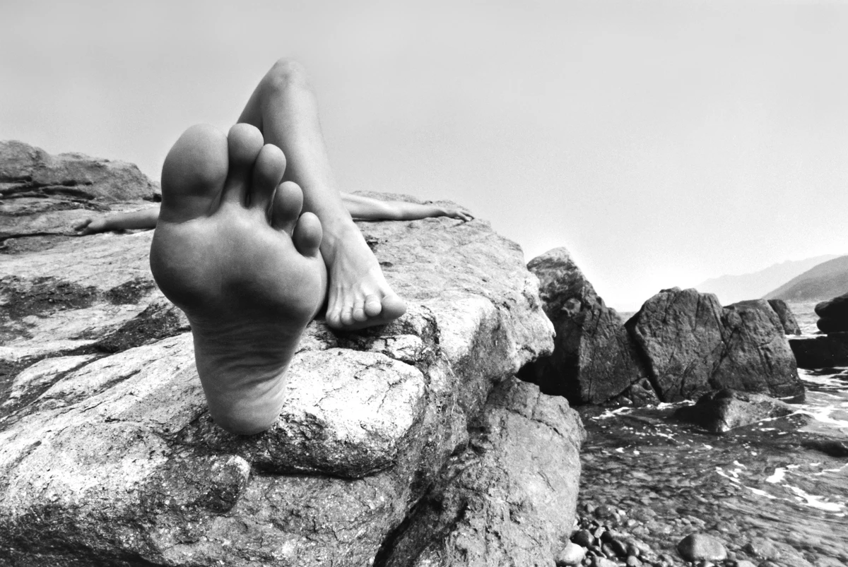 Black and white fine art nude photograph of a reclining female figure on coastal rocks, with an oversized foot in the foreground and the sea and distant mountains beyond.