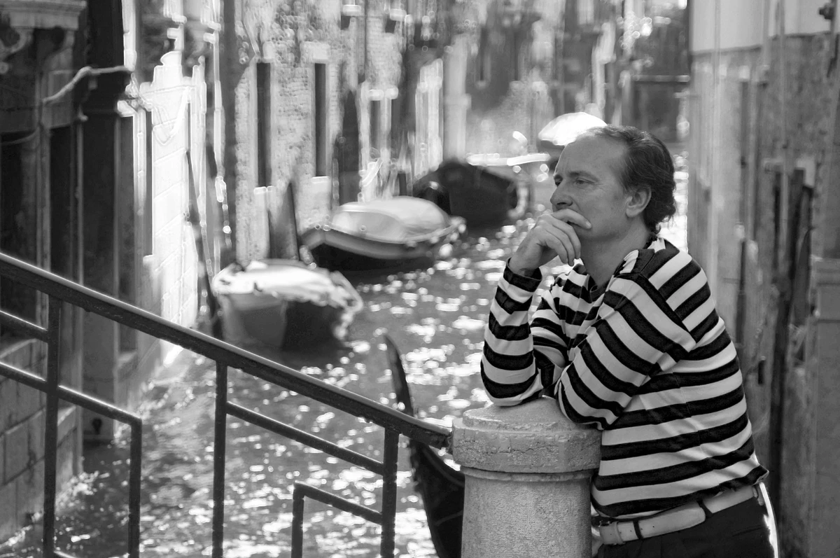 Man in a striped shirt leaning on a bridge overlooking a Venetian canal, with moored boats beyond in black and white.